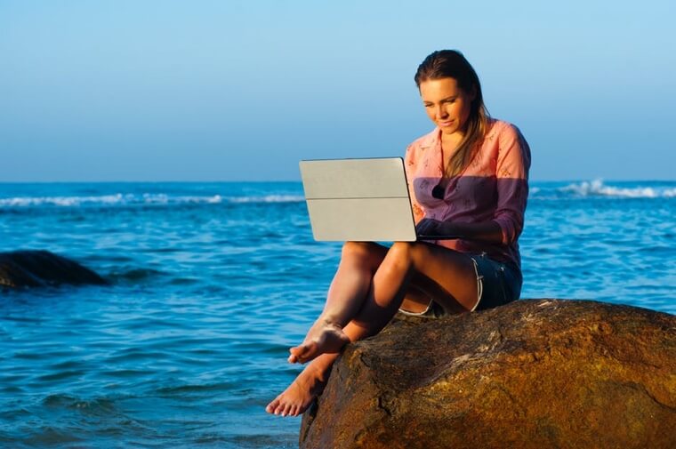 Woman typing on a laptop while sitting on a rock in the ocean