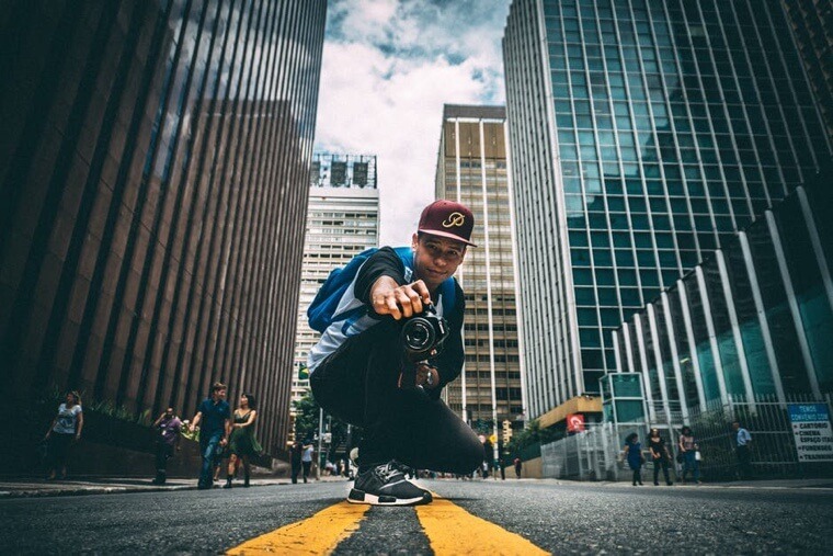 Man wearing a baseball cap crouched down in the middle of a street pointing a camera at the viewer