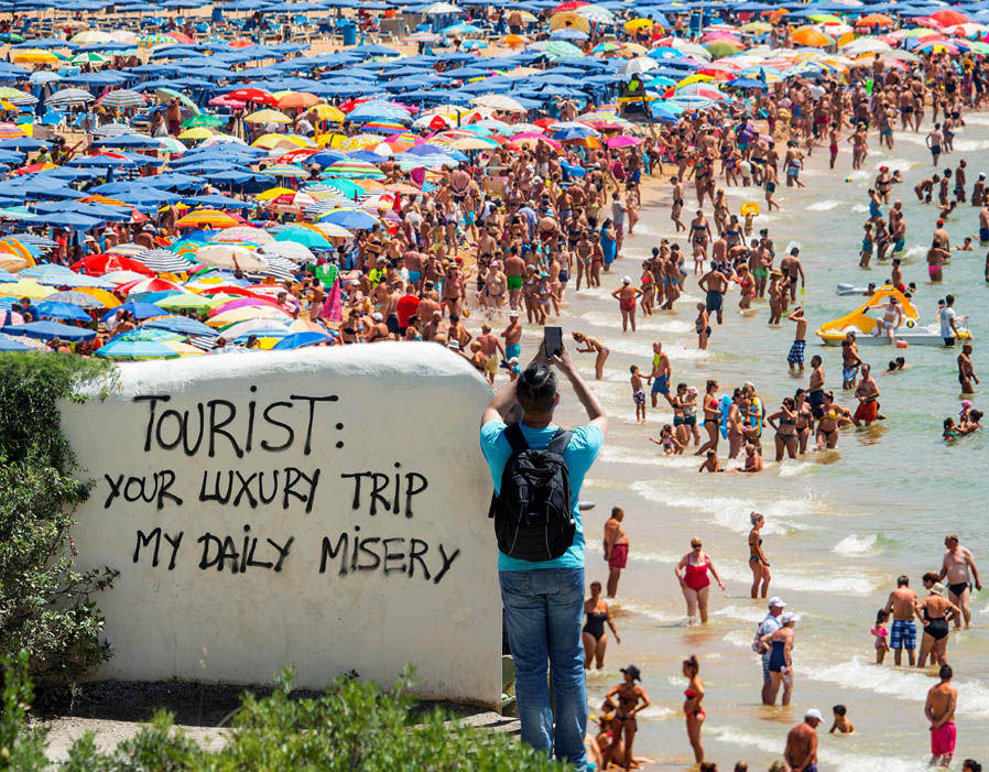 Overcrowded beach with a sign saying 'Tourist: your luxury trip, my daily misery'