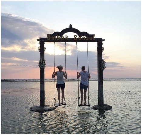 Two women standing on swings located on the ocean, swingset titled Ombak Sunset